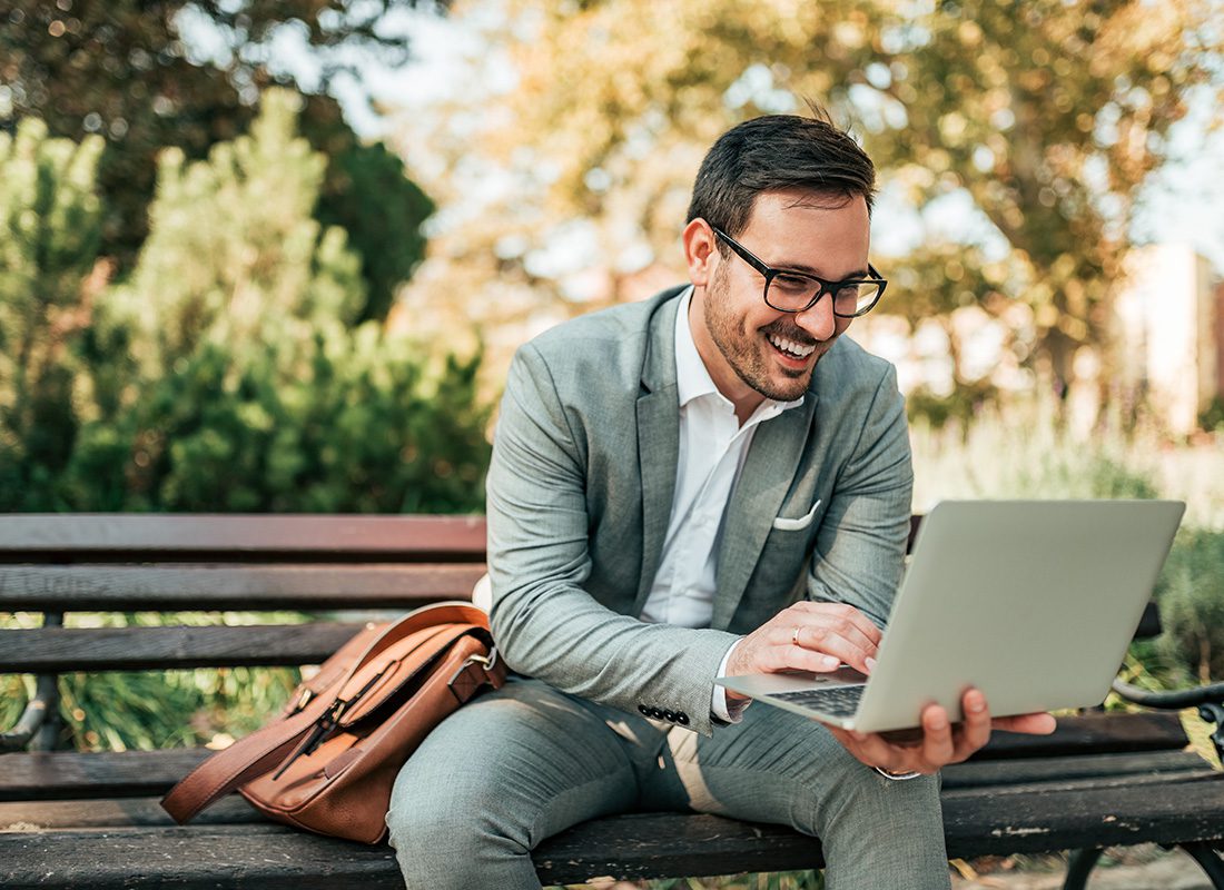 Blog - Professionally Dressed Man Smiling While Holding Laptop and Sitting on a Bench Outside in a Park on a Nice Day